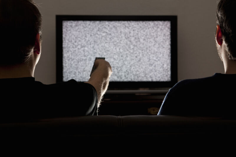 Two men sit in front of a TV that displays only static.