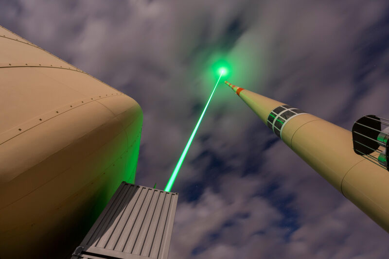 A green beam passes next to a thin tower beneath a partly cloudy sky.