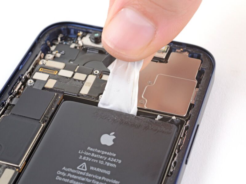 iPhone 12 battery being removed by fingers, with white adhesive stretching out beneath battery