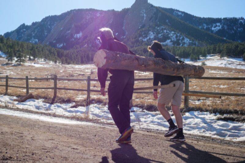 James Wilson and Rodger Kram carry a log with tumplines with the Boulder Foothills in the background.