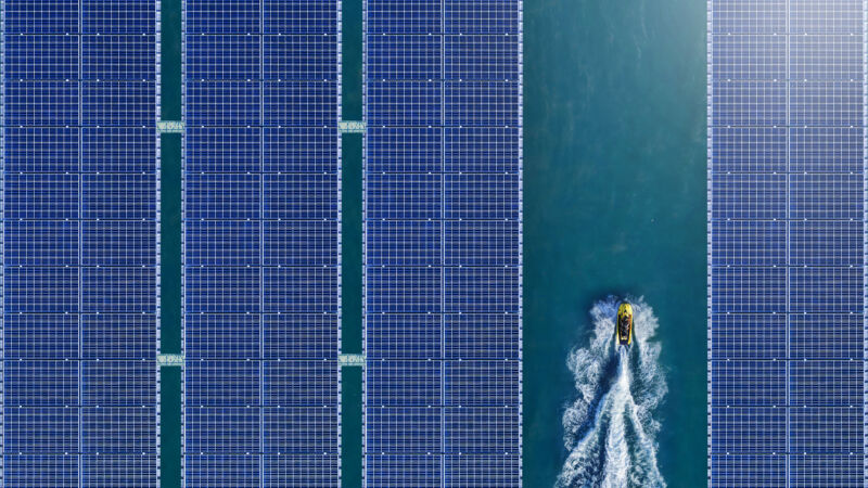 A vista aérea de uma represa com quatro fileiras de painéis solares instalados. Há um pequeno barco amarelo cruzando o vão entre as fileiras três e quatro.