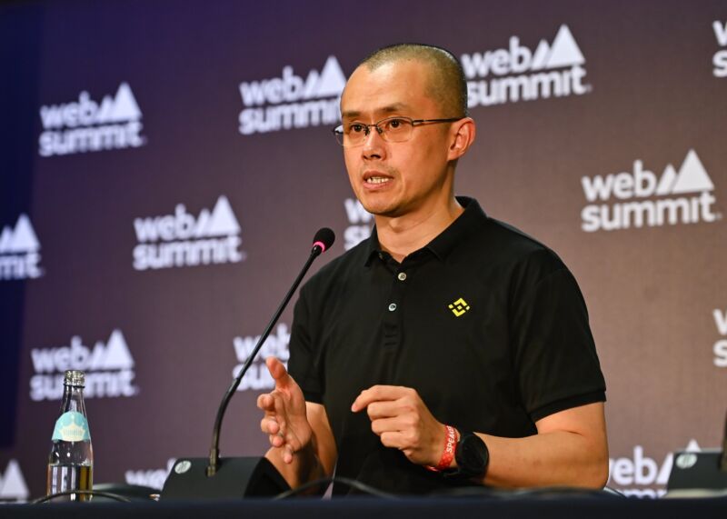 Binance founder and CEO Changpeng Zhao sits at a table and speaks into a microphone, wearing a shirt with a Binance logo.