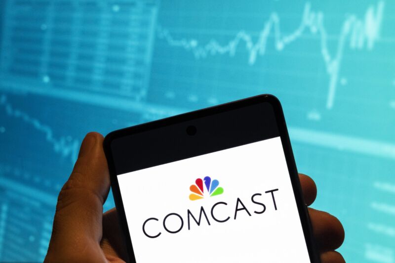 In this photo illustration, the Comcast logo is displayed on a smartphone with a stock exchange index graph in the background.