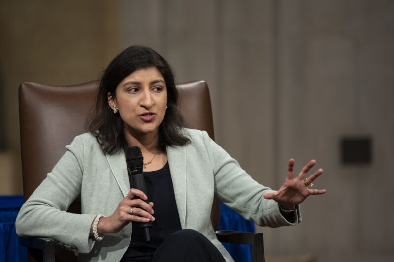 FTC Chair Lina Khan sitting in a chair and holding a microphone while she speaks at a conference.