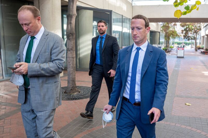 Mark Zuckerberg walks away from a courthouse while wearing a suit; he carries a phone in his left hand and a mask in his right hand.