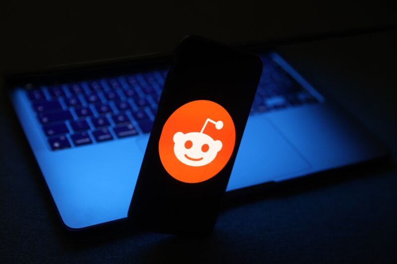 The Reddit logo displayed on a smartphone; a laptop is seen in the photo's background.