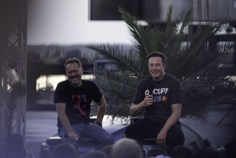SpaceX CEO Elon Musk and T-Mobile CEO Mike Sievert sit on a stage outdoors, holding microphones and smiling, during a press conference. Sievert wears a T-Mobile T-shirt and Musk wears a T-shirt that says 