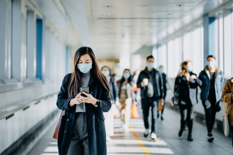 Asian woman with protective face mask using smartphone while commuting in the urban bridge in city against crowd of people