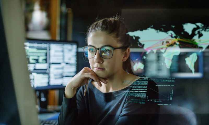 Imagen de archivo de una mujer joven, con anteojos, rodeada de monitores de computadora en una oficina oscura.  Frente a ella hay una pantalla transparente que muestra un mapa del mundo con algunos datos.