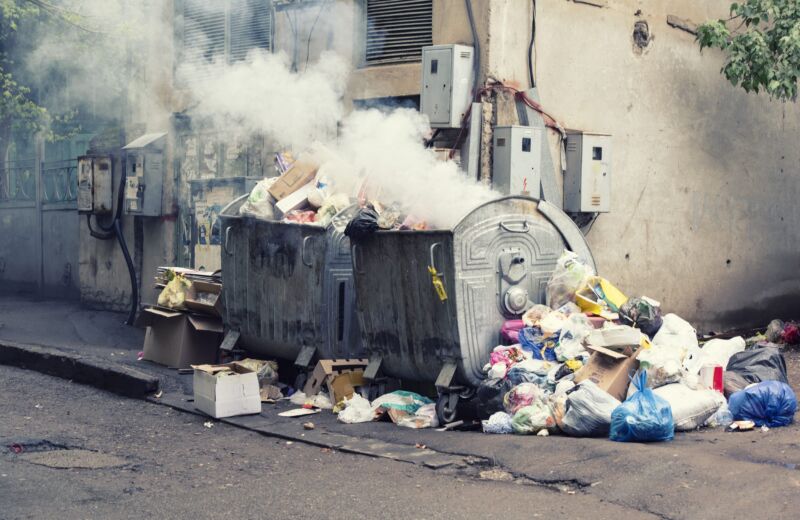 Smoke billows out of a burning trash dumpster. Trash bags are piled up on the sidewalk next to the dumpster.