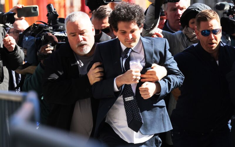 FTX Founder Sam Bankman-Fried arrives at court, surrounded by photographers and other people. One man appears to be holding Bankman-Fried around the torso and escorting him.