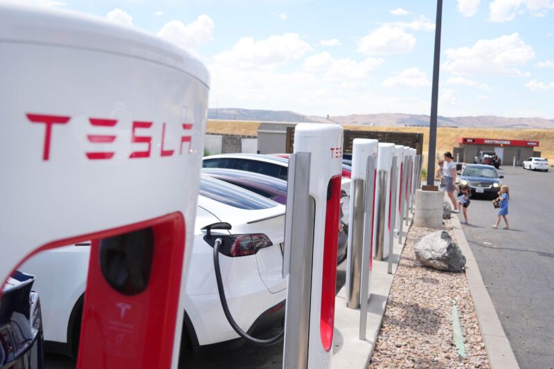 A line of electric cars connected to Tesla chargers in a parking lot outside during daytime.