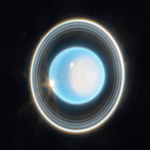 Planet Uranus on a black background.  The planet appears light blue, with a large white spot on the right side.  At the edge of that patch in the upper left is a bright white spot.  There is another white spot on the left side of the planet at 9 o'clock. Around the planet is a system of overlapping rings.  Uranus rings are vertical.