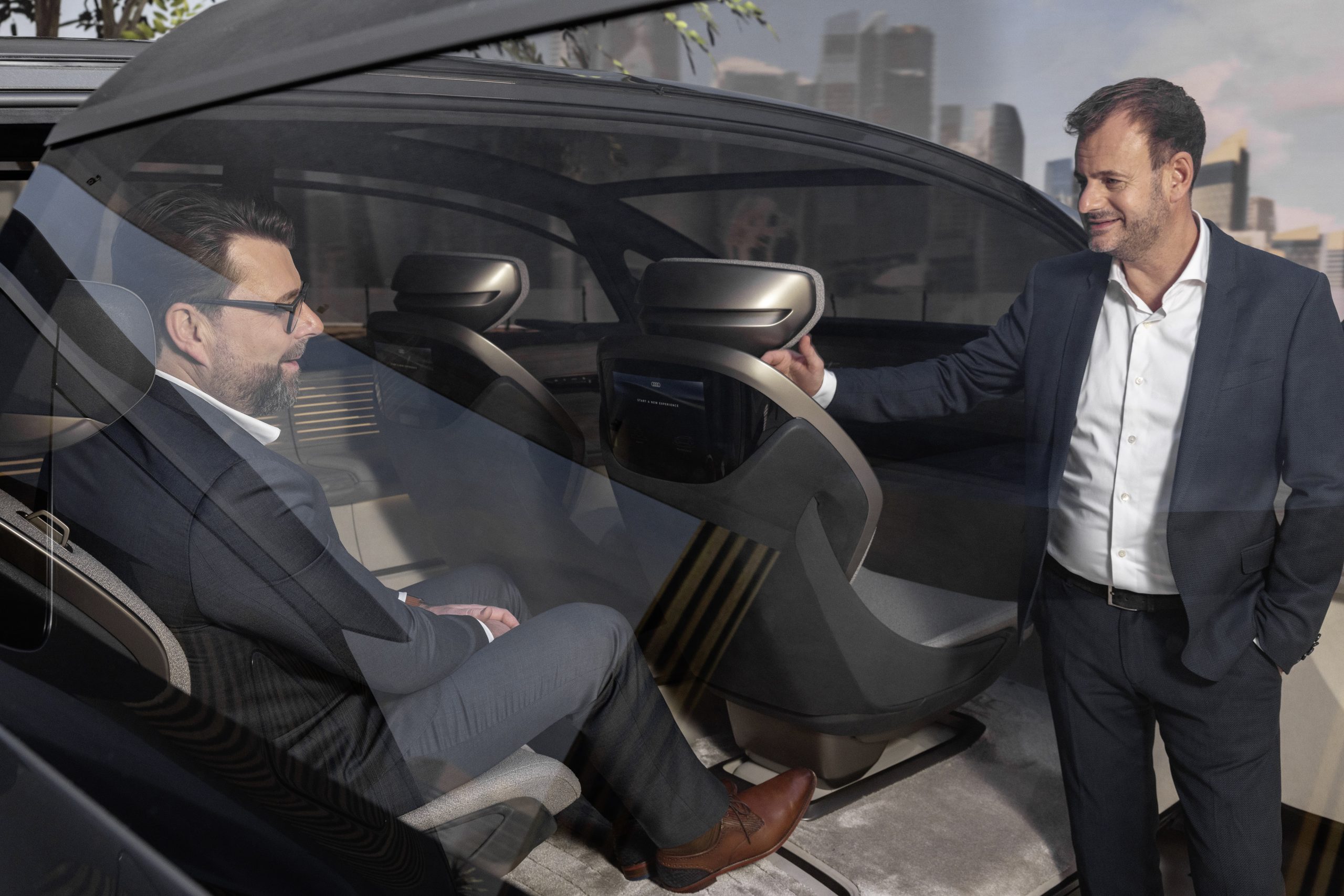A man sits in a concept car interior and talks to a man standing outside the car