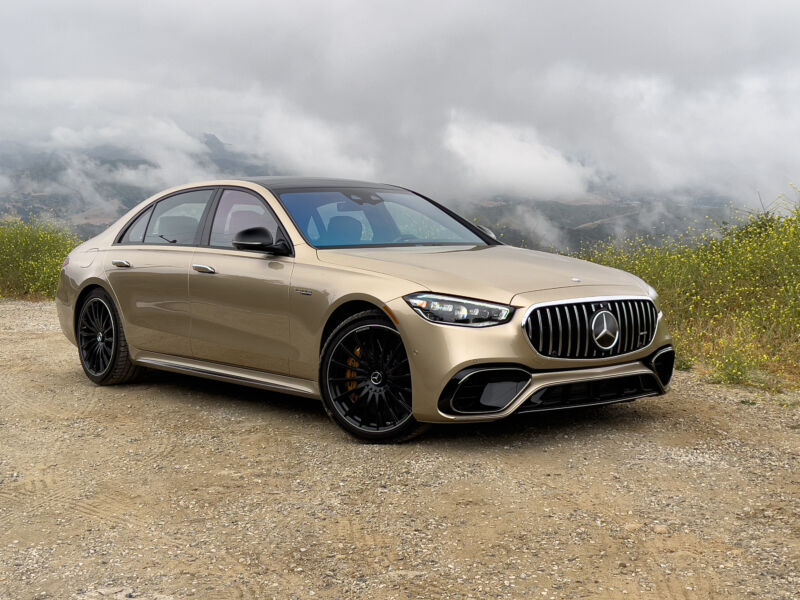 A gold mercedes sedan parked on a mountain road with clouds in the background
