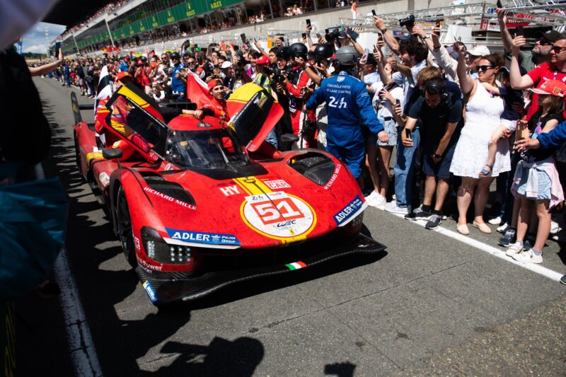 A red race car moves slowly down the pitlane at Le Mans after the race. There are hundreds of people cheering it on