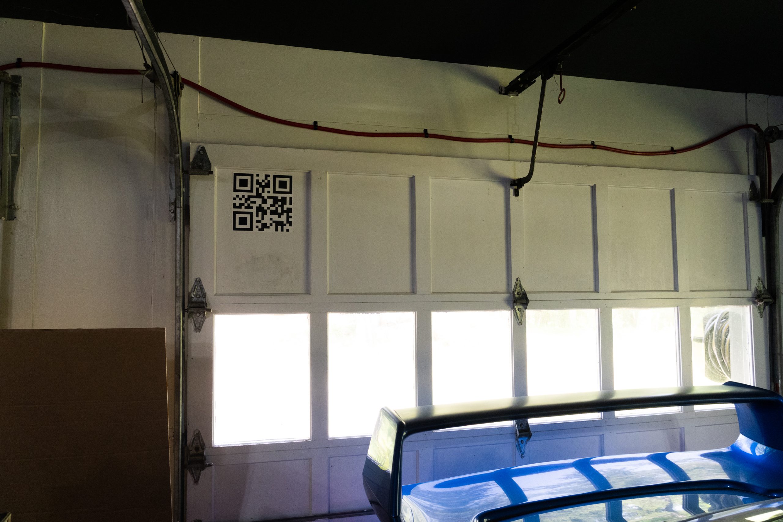 A white garage door with a QR code in the upper left corner. In front of it, the rear wing of a blue Subaru STI