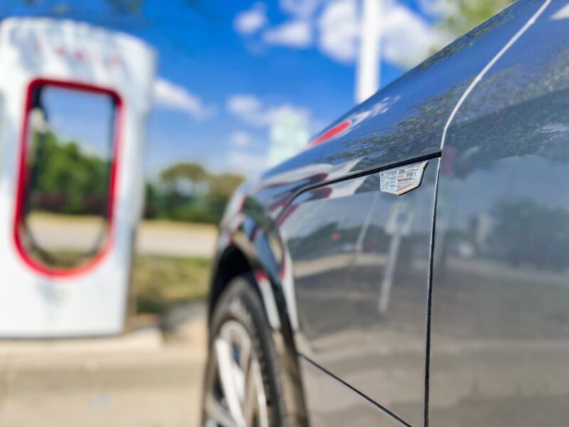 A close-up view of the front fender badge and charge door on a Cadillac LYRIQ parked at a Tesla Supercharger station.