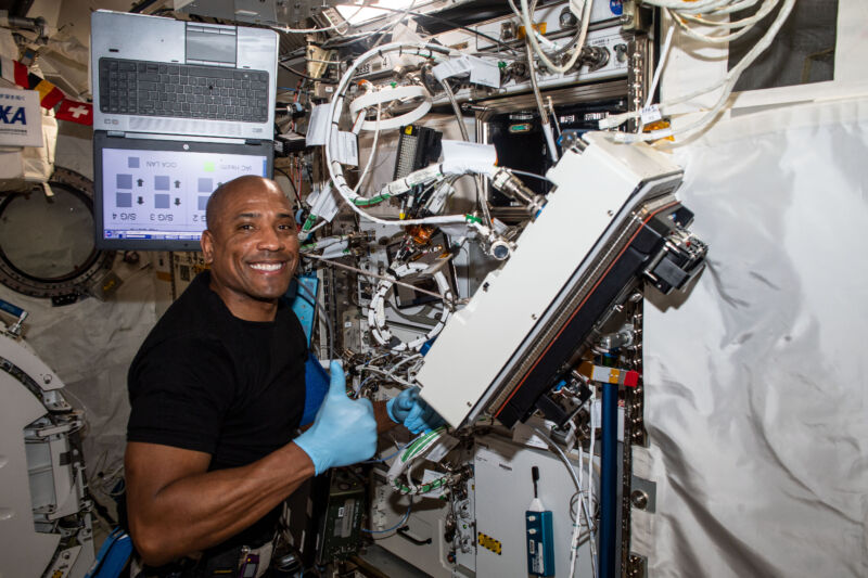 An astronaut stands in front of a complicated rack of equipment.