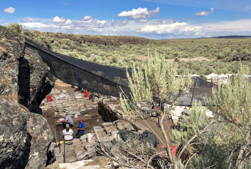 photo of a low rock cliff with black netting stretching from its top down to scrub desert landscape below, with archaeologists working beneath the netting in the shade