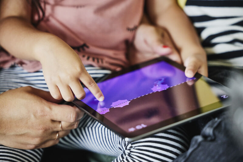 Close up shot of young girls hands playing game on digital tablet while sitting in parents lap