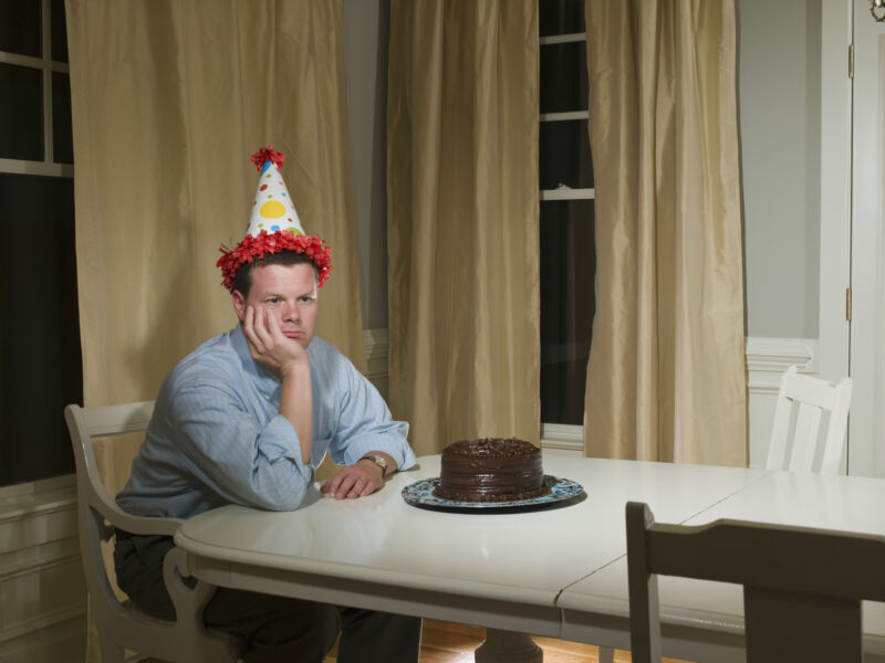 A person sitting alone at a table with a cake on it. The man is wearing a festive hat.