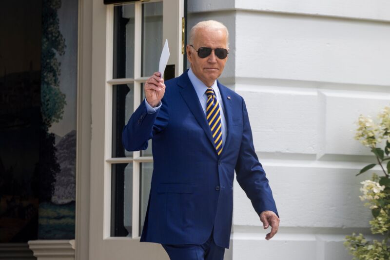 Joe Biden walking outside the White House, wearing sunglasses and holding a stack of index cards in his right hand.