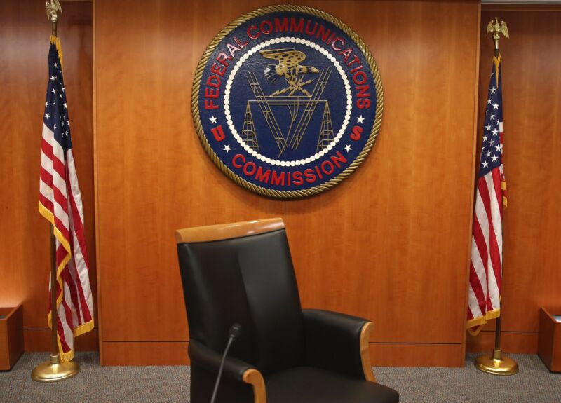 In the FCC hearing room, an empty chair sits in front of the FCC seal and two US flags.