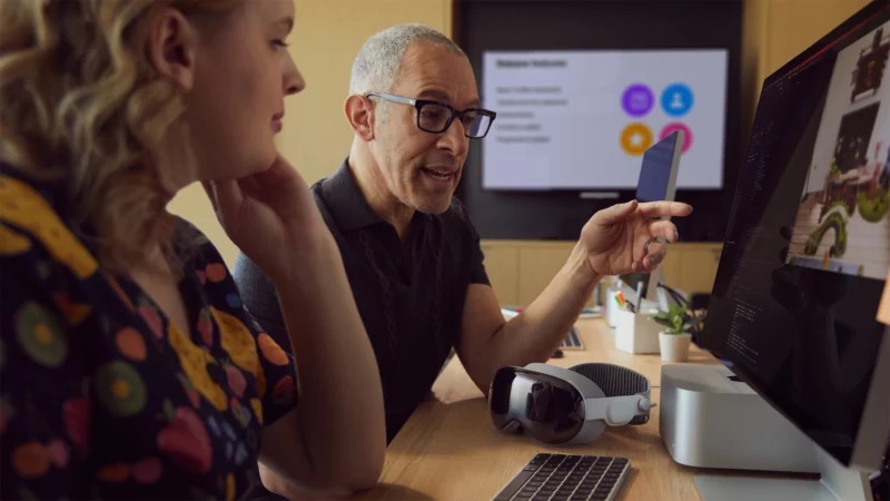 Los desarrolladores se sientan frente a una Mac con un auricular Vision Pro sobre la mesa