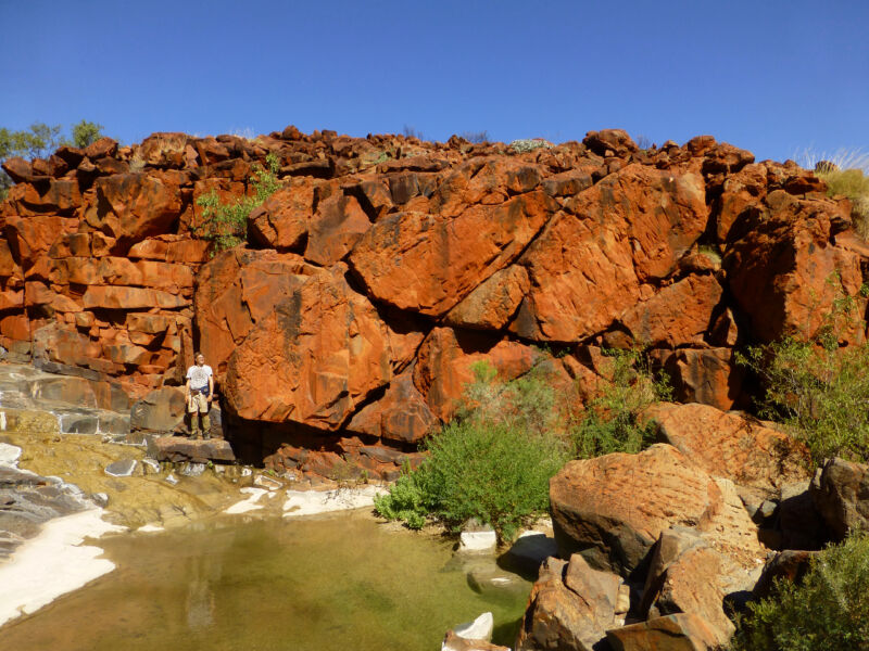 Image of a small person standing in front of large, reddish rocks.