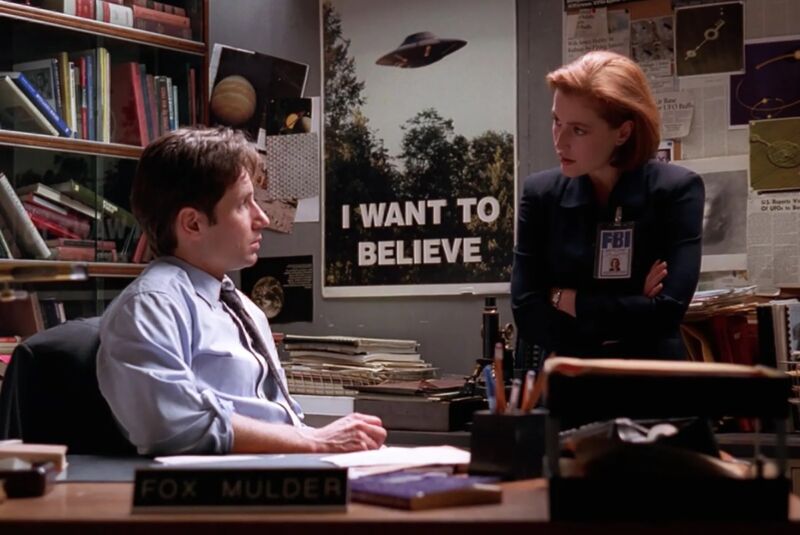 Mulder sitting at his desk, Scully sitting on top of it, with 