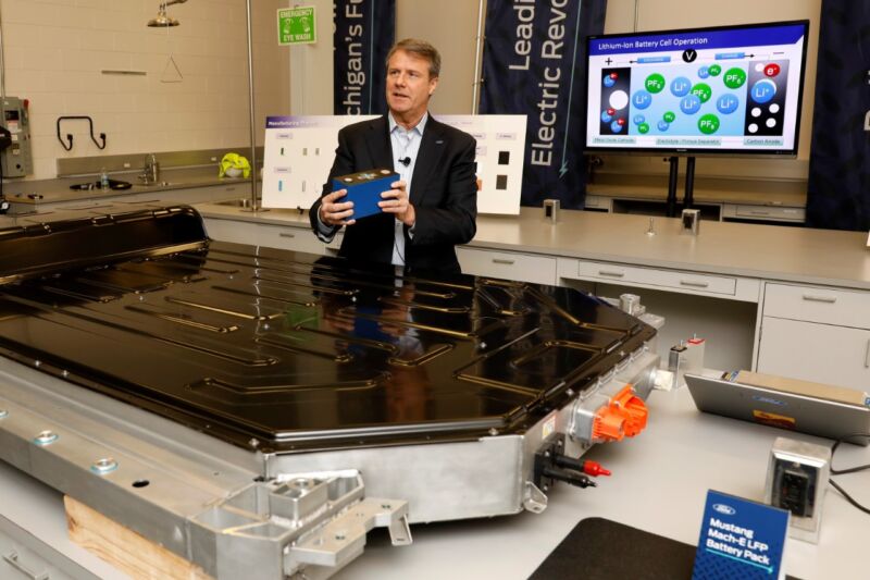 A man stands next to an EV battery with some posters in the background.