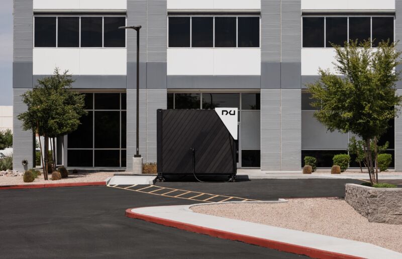 A black cube with a white triangle on its corner sits in front of an office building. The cube is the Nxu One charger.