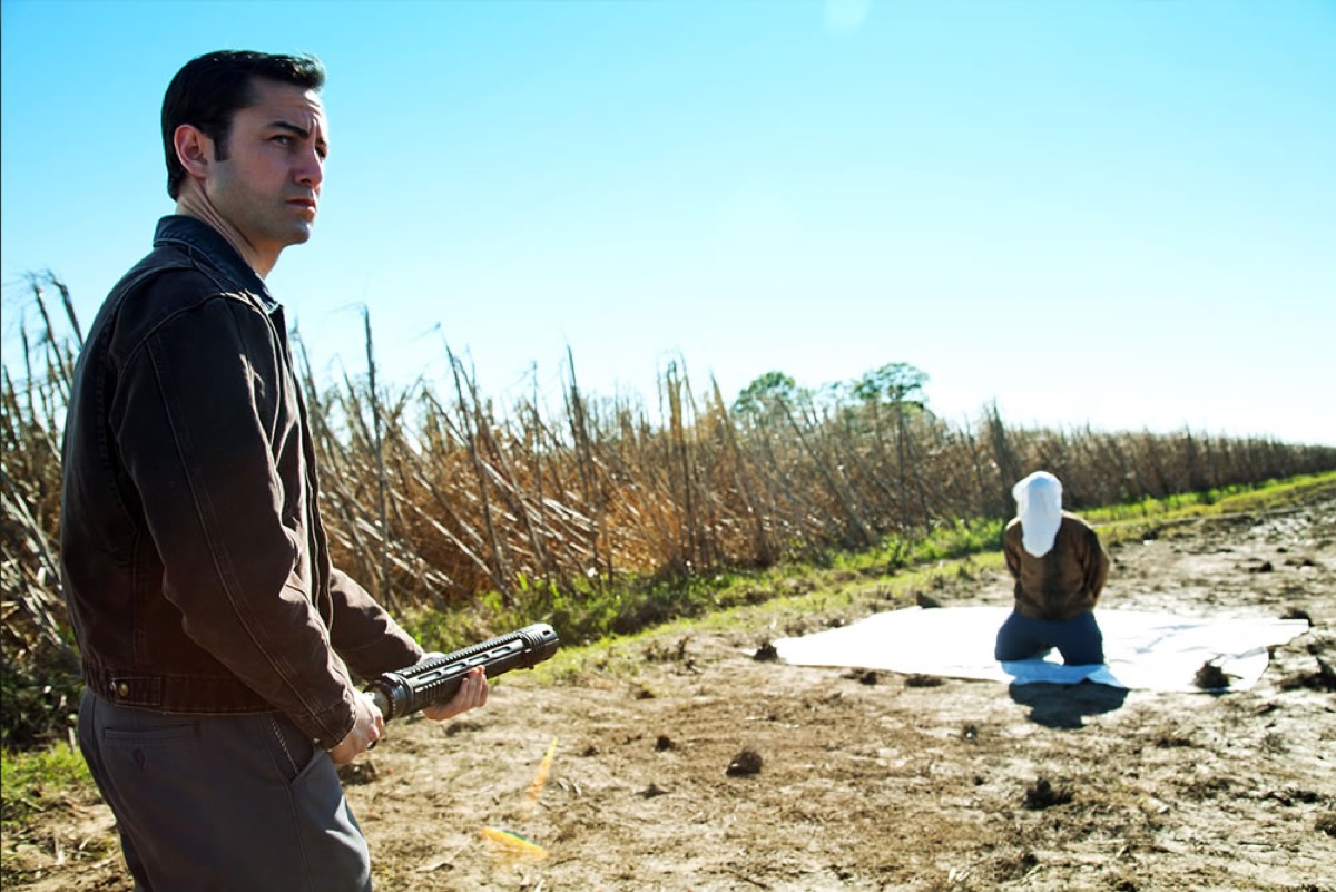 young man with gun standing in front of hooded man on his knees in a field