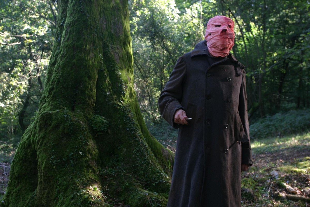 strange man in long black coat with face bandaged in pink wraps standing next to a tree