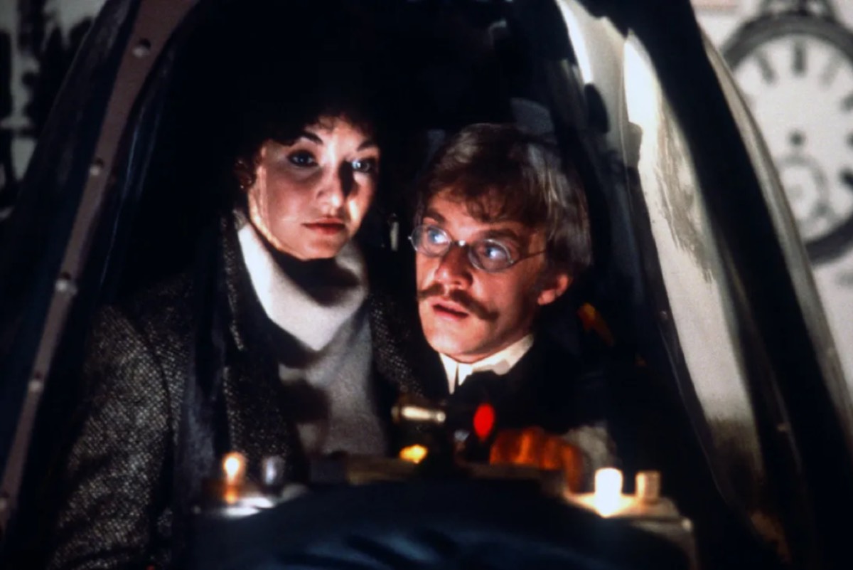 1970s woman and late 19th century man sitting close together in a pod-like time machine
