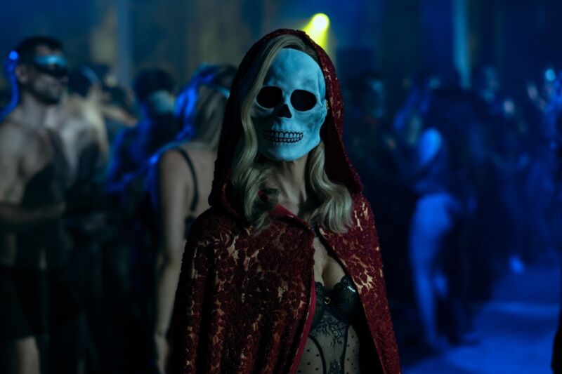 mysterious female figure in red cape and skull mask staring directly into the camera while revelers dance behind her
