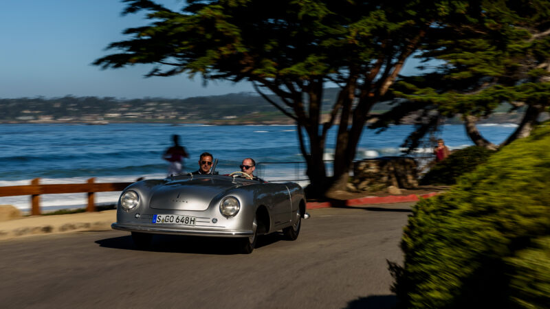 Two people drive in a silver open-top car by the sea with a big tree in the background