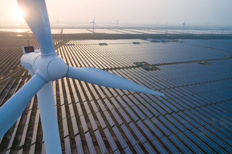 Wind turbines stand above a large field of solar panels in a view backlit by the rising sun.