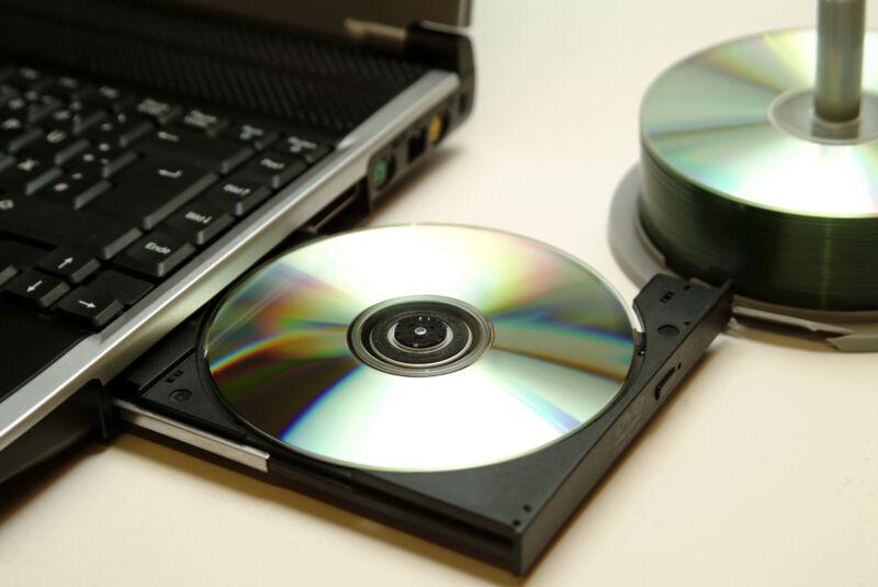 Blank CD inserted into a laptop CD drive, with a spindle of blank CDs nearby.