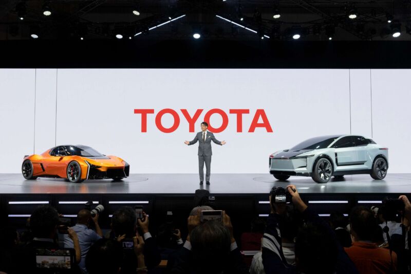 A man stands on stage with a concept car either side of him. In the background it says TOYOTA in large red letters