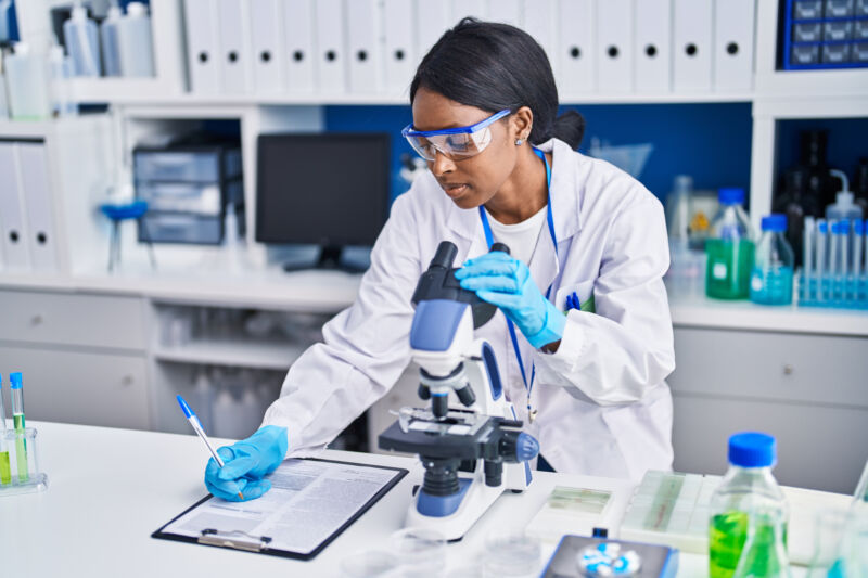 A woman adjusts a microscope while taking notes on a clipboard.