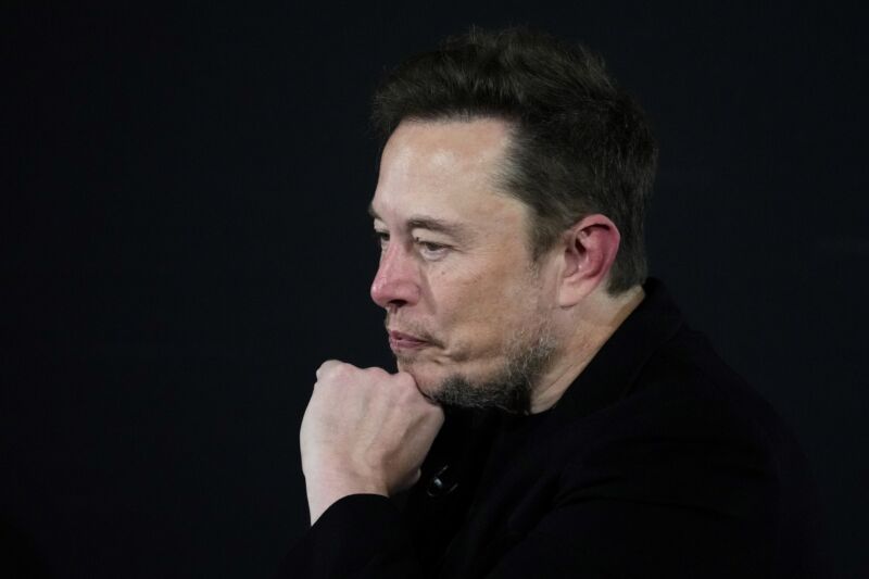 Elon Musk on stage at an event, resting his chin on his hand