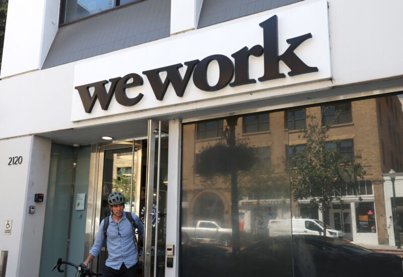 A man wearing a bicycle helmet walks out of a building that has a WeWork logo on its front.