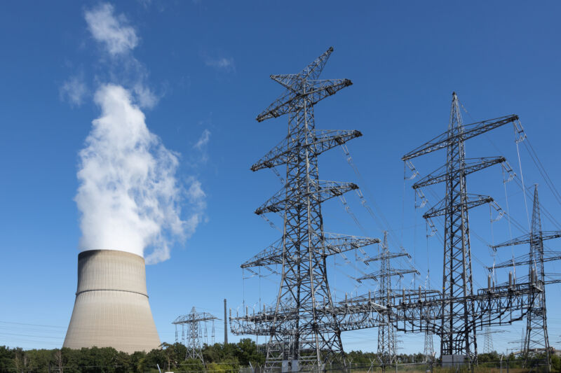 Imagen de líneas eléctricas con una torre de enfriamiento de una central eléctrica al fondo.