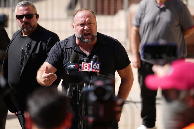 Alex Jones speaking outside a court house while standing in front of several TV news microphones.