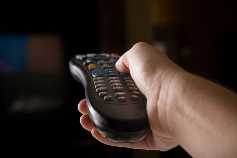 A hand pointing a TV remote control at a TV against a dark background.