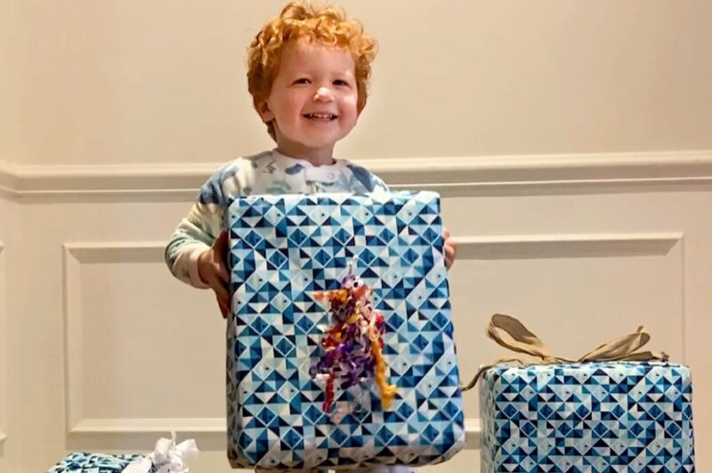 adorable curly red haired toddler in onesie grinning while holding a wrapped christmas present