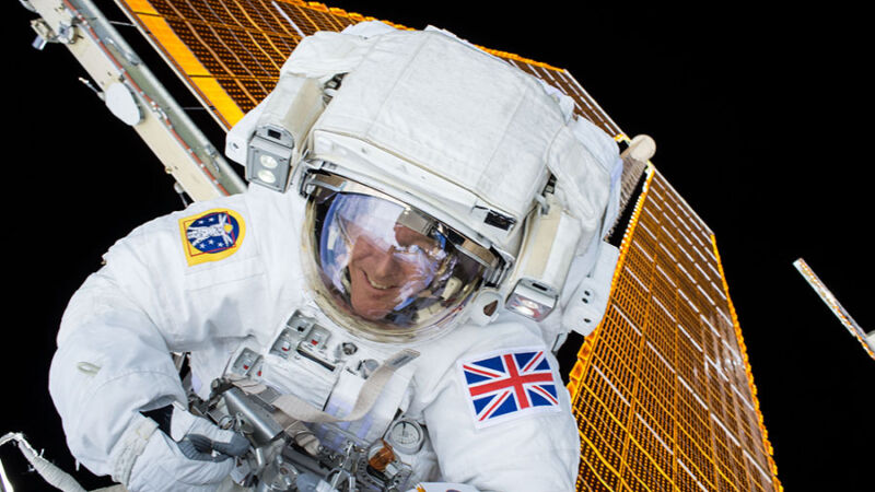 Image of a smiling person inside a spacesuit, with a solar panel and the blackness of space behind him.