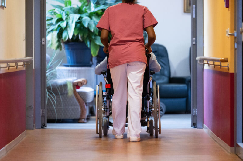 A nursing home resident is pushed along a corridor by a nurse.